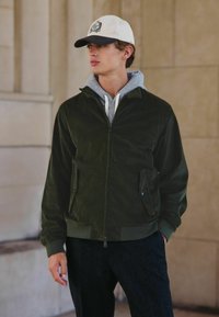 Olijfgroene corduroy jasje met een ritssluiting aan de voorkant, zijzakken en een grijze hoodie eronder. Gepaard met donkere gestructureerde broek en een pet.