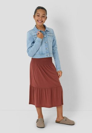 Meisje dat glimlacht en staat, draagt een lichtblauwe spijkerjack, een roestkleurige midirok en beige instapschoenen tegen een effen achtergrond.