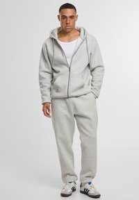 Jasnoszara bluza zip-up z dużymi przednimi kieszeniami i miękką strukturą, w komplecie z pasującymi spodniami dresowymi. Biała koszulka na ramiączkach pod spodem.