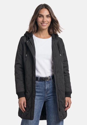 Femme portant une veste matelassée noire à capuche par-dessus une chemise blanche et un jean bleu, souriante devant un fond uni.