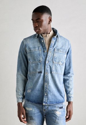 Lichtblauw denim overhemd met twee voorzakken, versleten details en knoopsluiting. Draagt over een beige top met hoge kraag.