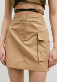 Beiger Baumwollrock mit Seitentasche, strukturiertem Design und geradem Saum. Glatte Textur; minimalistischer Stil ohne zusätzliche Muster.