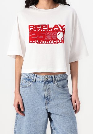Kobieta w białym krótkim T-shircie z czerwonym tekstem "Replay Montecarlo 25 Country Club" i jasnoniebieskich, wysokich dżinsach.