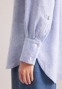 Camicia azzurra a righe con strisce bianche, dotata di polsini con due bottoni e orlo arrotondato, realizzata in un tessuto testurizzato.