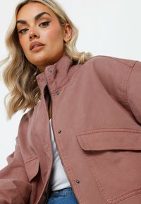 Rosa kordjacka med hög krage, framdragkedja, tryckknappar och två bröstfickor, som visar en mjuk textur och en strukturerad design.