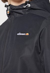 Czarna nylonowa kurtka o fakturowanej powierzchni, z zamkiem błyskawicznym z przodu i kapturem. Logo marki znajduje się na lewej piersi.