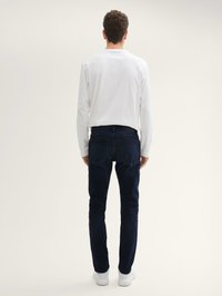 Camiseta blanca de manga larga combinada con jeans de color azul oscuro. Diseño ajustado, textura suave y detalles en el bolsillo trasero. El calzado son zapatillas blancas.