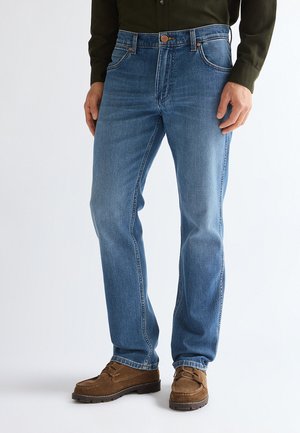Homme portant un jean bleu droit, des chaussures en daim marron et une chemise à manches longues vert foncé, debout sur un fond blanc.