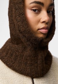 Brązowy dzianinowy kołnierz wykonany z teksturowanej wełny, charakteryzujący się grubym wzorem ściągacza. Otacza szyję i częściowo zasłania twarz.