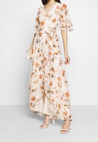 Robe maxi en tissu rose clair avec un motif floral orange et vert. Caractéristiques d'un col en V, de manches courtes et d'un design enveloppant. Talons blancs et une pochette.