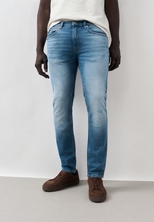 Mężczyzna w jasnoniebieskich obcisłych dżinsach i brązowych trampkach, stojący na tle gładkiej jasnoszarej ściany i podłogi.