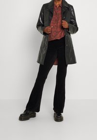 Svarta corduroy byxor med utfläkt ben, en rutiga röd och grå skjorta, och en glänsande mörkgrön trenchcoat. Svarta plattformsskor fulländar looken.