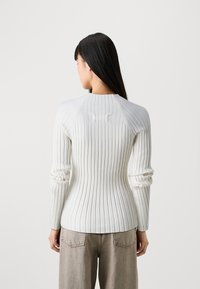 Enganliegendes weißes Rippoberteil mit langen Ärmeln, Rundhalsausschnitt und subtiler Textur, kombiniert mit hellbeigen, hochgeschnittenen Jeans.