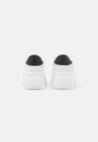 Zapatillas blancas con una suela blanca texturizada y detalles en cuero negro en el talón. Material suave y diseño redondeado con un detalle mínimo.
