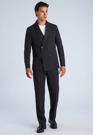 Mężczyzna stojący, ubrany w czarny dwurzędowy garnitur, czarne eleganckie buty i białą koszulę pod marynarką na jednolitym tle.