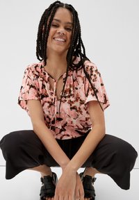 Bluse mit floralem Pinkdruck und kurzen Ärmeln, mit Schleifen-Detail am Ausschnitt, kombiniert mit lockeren schwarzen Hosen und geflochtenen Sandalen.