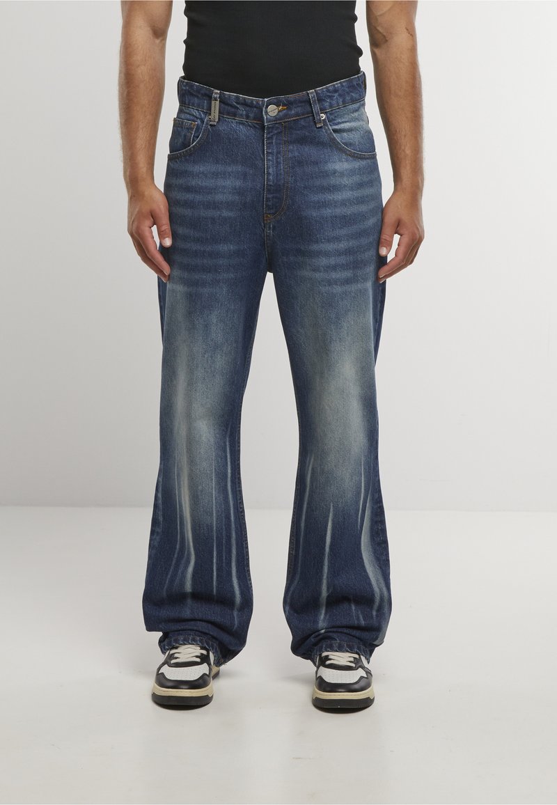 Homme portant un jean bleu délavé et ample avec des baskets, debout contre un fond clair uni.