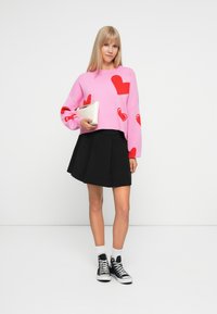 Pink sweater with red heart patterns, black pleated skirt, and black high-top sneakers. Holding a white clutch. Smooth and casual design.