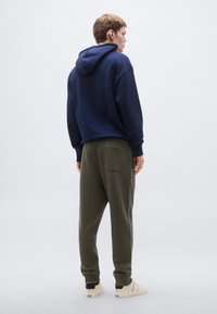 Tengerészkék kapucnis pulóver húzózsinórral, olívazöld színű melegítőnadrággal, amelynek hátul zsebe van. Világos színű cipővel egészül ki a szett.