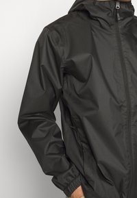 Veste imperméable noire avec fermeture éclair à l'avant, poignets élastiques, capuche et poches latérales, fabriquée en matériau lisse et léger.