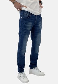Jeans de mezclilla azul oscuro con un ajuste slim, un ligero desgastado, cremallera y diseño de cinco bolsillos, combinados con zapatillas blancas.