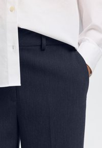 Persona che indossa pantaloni eleganti blu scuro con la mano in tasca e una camicia bianca a maniche lunghe infilata dentro.