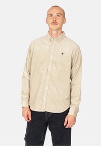 Camisa de botones de sarga beige con cuello y un pequeño logo negro, combinada con jeans oscuros. Presenta una superficie texturizada y mangas largas.