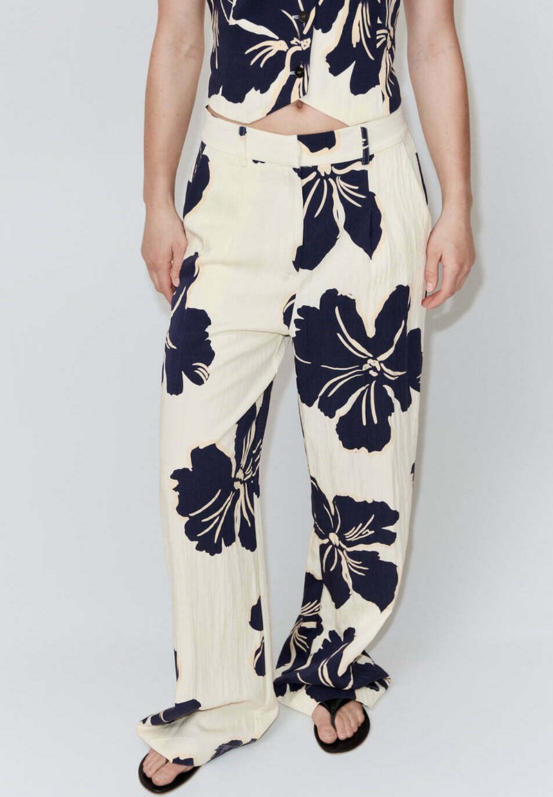 Femme portant un pantalon large couleur crème avec de grands motifs floraux foncés et un top court boutonné assorti, porté avec des sandales noires.