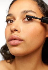 Czarna szczoteczka do tuszu do rzęs nakładana na wydłużone czarne rzęsy. Skóra jest gładka w naturalnym odcieniu, a usta są jasne i lekko błyszczące.