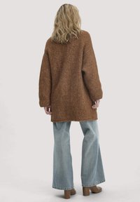 Person mit welligem, blonden Haar, der einen langen, braunen Strickpullover, hellblaue weite Jeans und braune Stiefeletten trägt, steht mit dem Rücken zur Kamera.