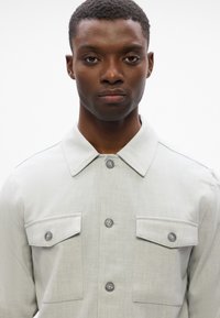 Ανοιχτό γκρι πουκάμισο με δομημένο γιακά, δύο τσέπες στο στήθος και κουμπιά μπροστά. Το υλικό έχει λεία υφή, κατάλληλο για καθημερινή χρήση.