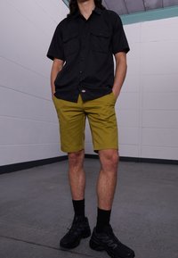 Černé tričko s krátkými rukávy a dvěma předními kapsami, které je nošeno s olivově zelenými kraťasy. Černé sportovní boty se tmavými ponožkami. Jednoduchý, neformální oděv.