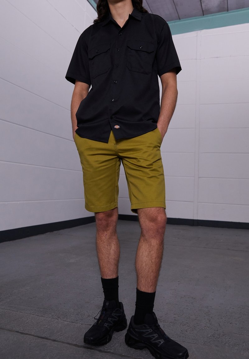 Černé tričko s krátkými rukávy a dvěma předními kapsami, které je nošeno s olivově zelenými kraťasy. Černé sportovní boty se tmavými ponožkami. Jednoduchý, neformální oděv.