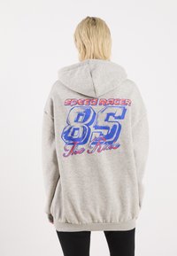 Grå hoodie med et stort print på ryggen, der præsenterer "SPEED RACER", "85" og "The Ride" i blå og rød grafisk skrift. Struktureret stof.