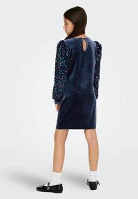 Granatowa aksamitna sukienka z długimi rękawami ozdobionymi cekinami oraz dekoltem w kształcie łezki z tyłu, zestawiona z czarnymi butami i białymi skarpetkami.