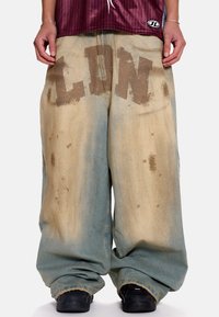 Φαρδιά τζιν παντελόνια σε ανοιχτό μπλε και μπεζ με εφέ πλυσίματος οξέος. Διαθέτουν μεγάλο, ξεθωριασμένο κείμενο "LDN" σε καφέ χρώμα στο εμπρός μέρος.