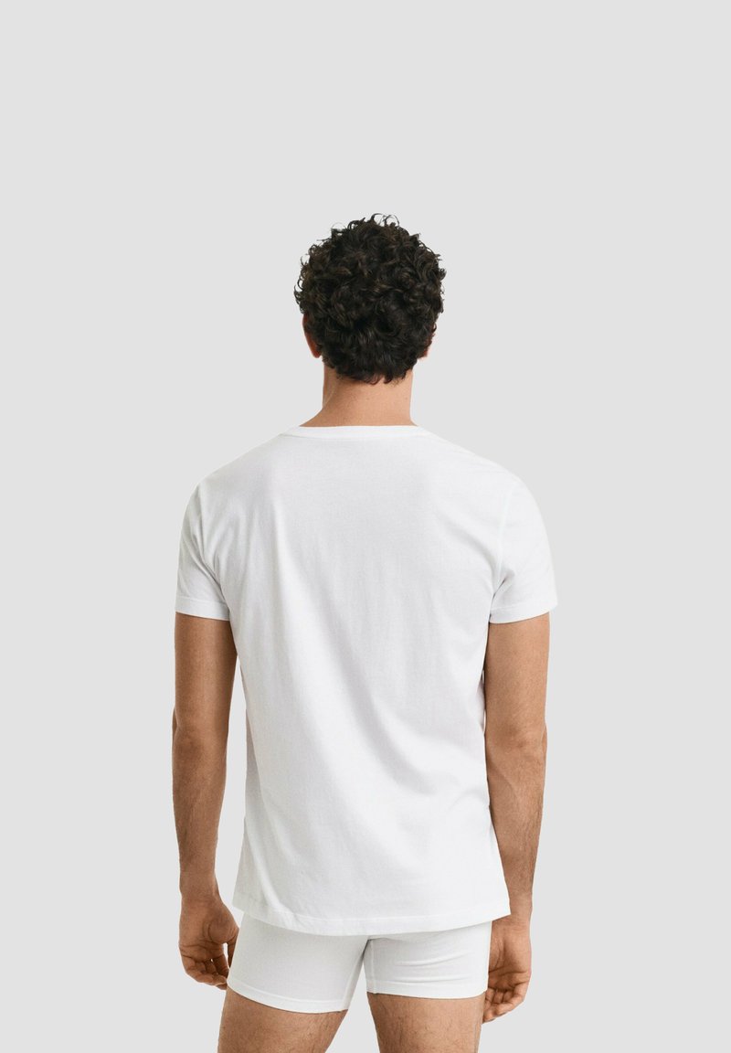 Uomo con capelli ricci scuri visto da dietro, che indossa una maglietta bianca semplice a maniche corte e boxer bianchi su sfondo grigio chiaro.