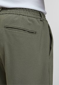 Pantalons de jogging vert olive avec taille élastique, dotés d'une poche latérale et d'une poche arrière avec un détail de doublure subtil. Texture lisse.