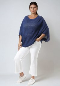 Marineblaues, offenes Strick-Poncho mit weiten Ärmeln, kombiniert mit weißen Hosen mit fransigem Saum und weißen Sneakers, was einen entspannten Look kreiert.