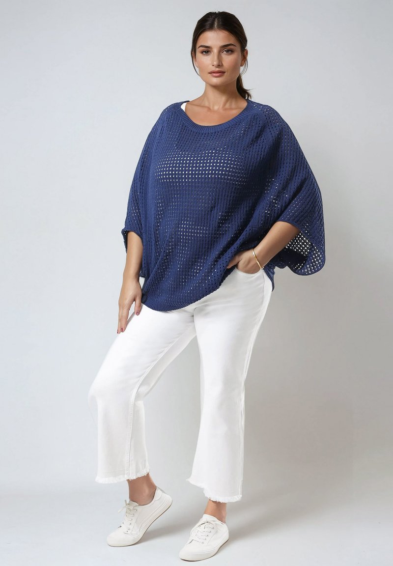 Marineblaues, offenes Strick-Poncho mit weiten Ärmeln, kombiniert mit weißen Hosen mit fransigem Saum und weißen Sneakers, was einen entspannten Look kreiert.