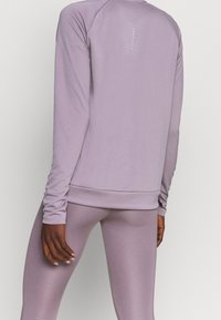 Helle lila Sportoberteil mit langen Ärmeln, gerippten Bündchen und Saum, auf dem Rücken mit vertikalen weißen Punkten versehen, kombiniert mit passenden Leggings.