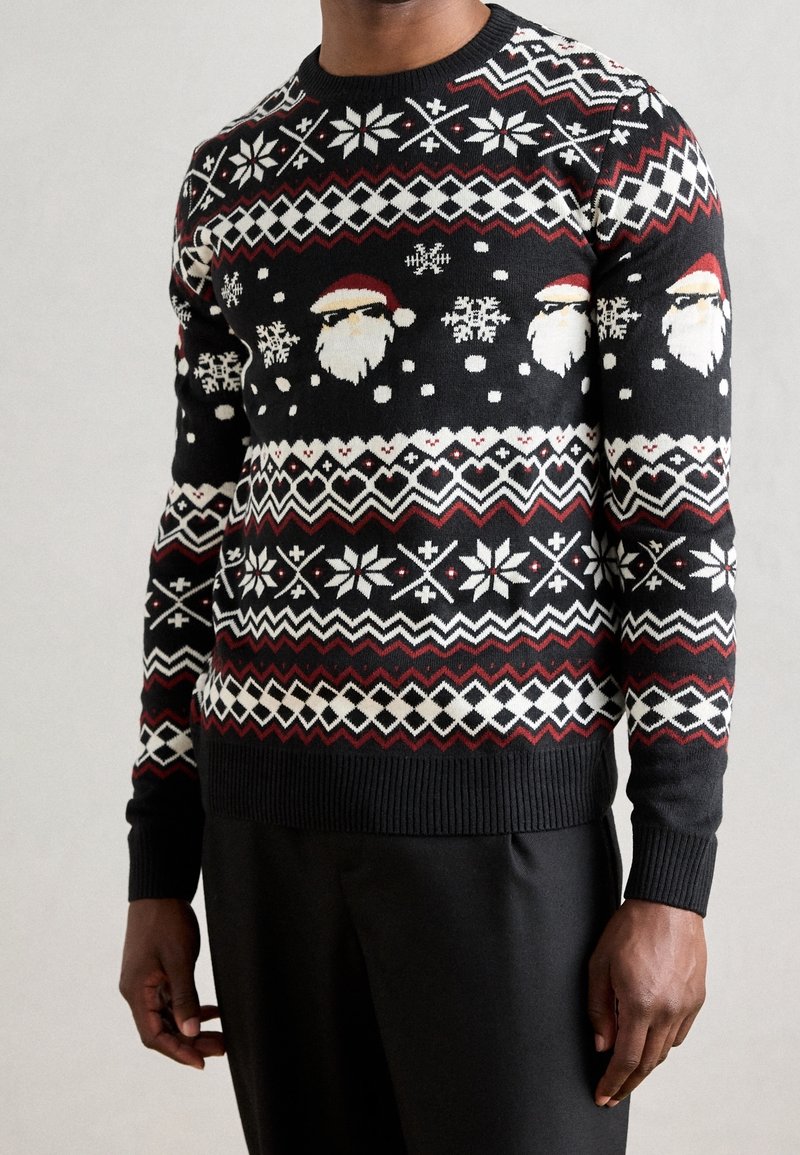 Pull de Noël noir avec des motifs géométriques blancs et rouges, des flocons de neige et des visages du Père Noël portant des lunettes de soleil sur une personne debout.