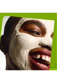 Närbild av ett leende ansikte med en lermask, mörk hy och en texturerad vit mask som täcker delar av ansiktet; livfull grön bakgrund som accent.