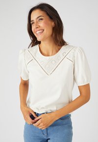 Blusa de algodón blanca con un yugo decorativo de encaje de ojetes con patrones florales, mangas cortas tipo globo y escote redondo.