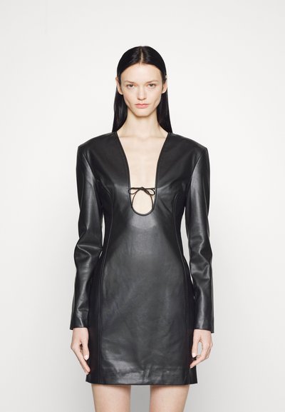 Žena oblečená v obtiahnutých čiernych kožených minišatách s dlhými rukávmi, silnými ramenami a hlbokým výstrihom do V, ktorý je stiahnutý malou mašľou.