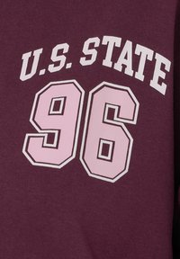 Felpa color bordeaux con testo bianco: "STATO USA" sopra grandi numeri rosa "96". Il tessuto ha una texture liscia con un design casual.