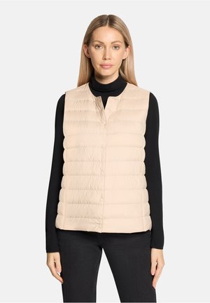 Vrouw draagt een beige gewatteerde mouwloze bodywarmer over een zwarte coltrui met lange mouwen en zwarte broek, staand tegen een effen achtergrond.