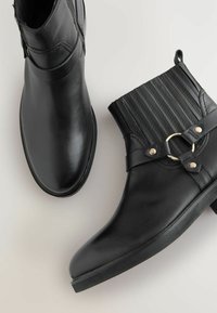 Botas de tobillo de piel negra que presentan un detalle de parte superior acanalada, un acento metálico circular y un tacón bajo apilado. Textura suave en todo momento.