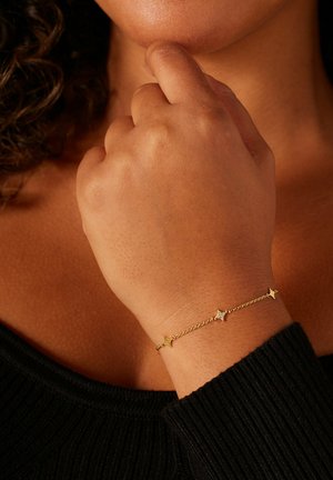 Bracelet en ton or avec des accents en forme d'étoile et de diamant sur une chaîne délicate, porté sur une main à la surface de peau texturée.