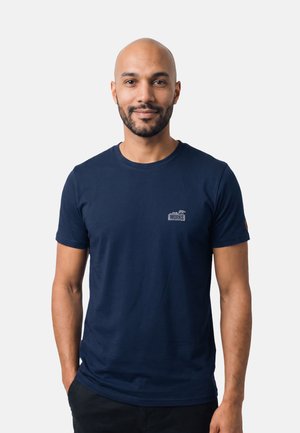 Uomo calvo sorridente con barba corta, che indossa una t-shirt blu navy e pantaloni neri, in piedi davanti a uno sfondo chiaro e uniforme.